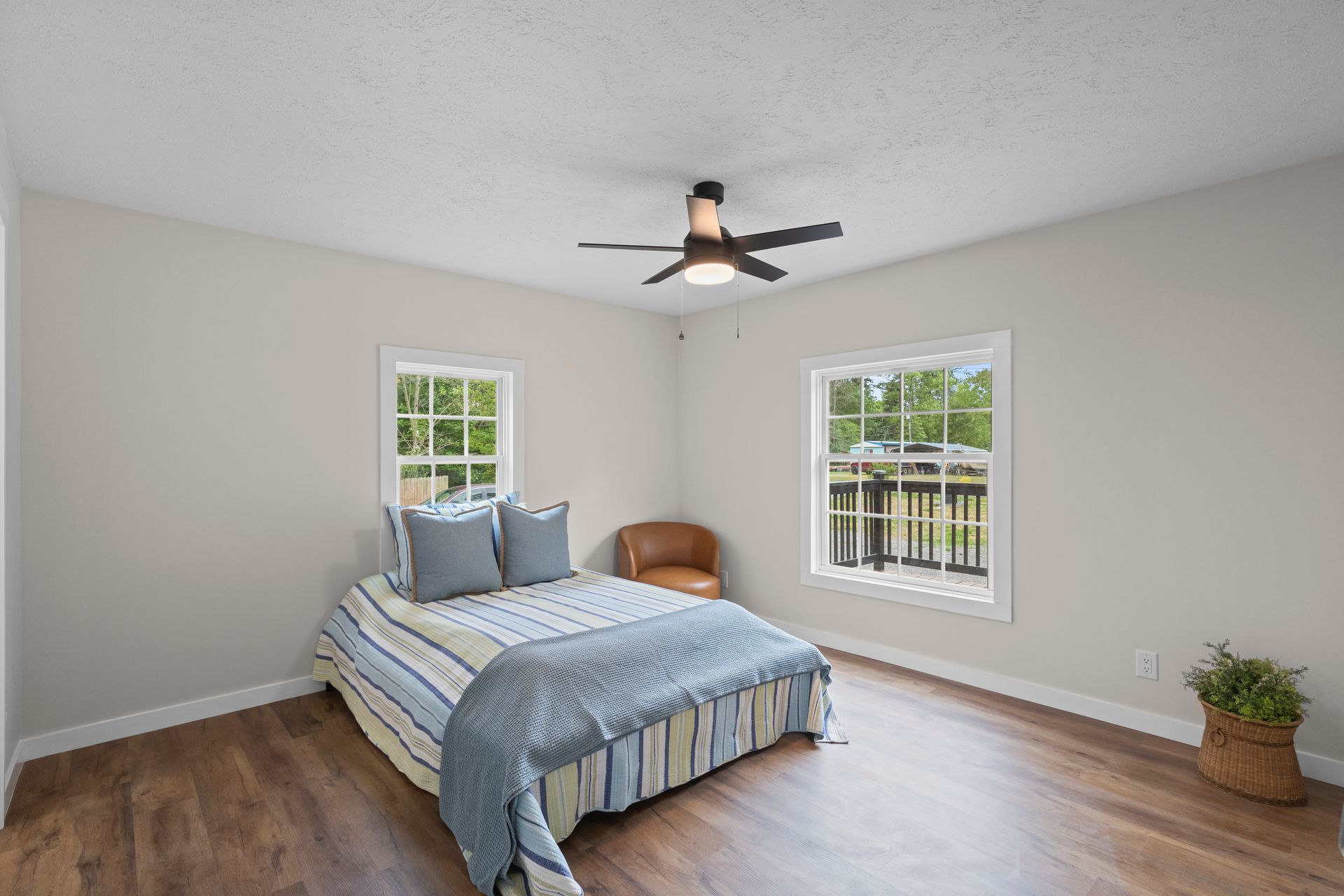 Bright bedroom with a bed, ceiling fan, two windows, wood floor, and a small plant in the corner