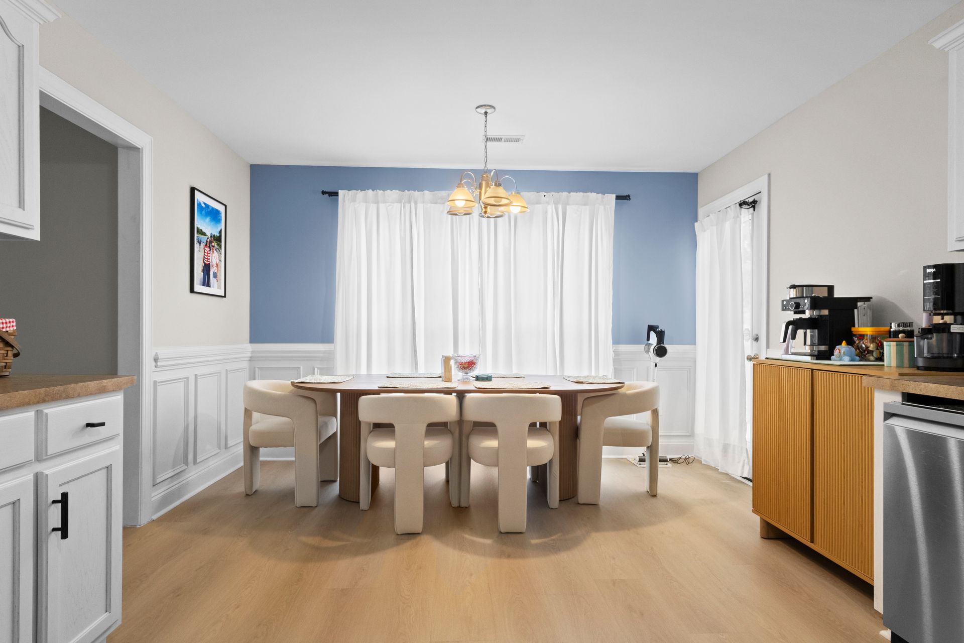 Bright dining room with round table, white chairs, blue accent wall, and sideboard near kitchen appliances