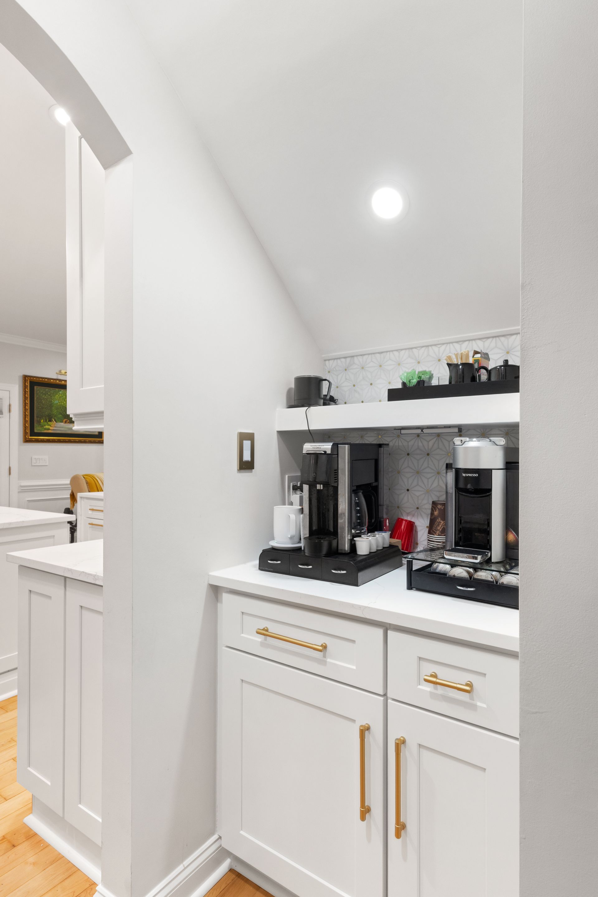 Narrow white kitchen nook with coffee machine, shelves, and brass handles under recessed ceiling light