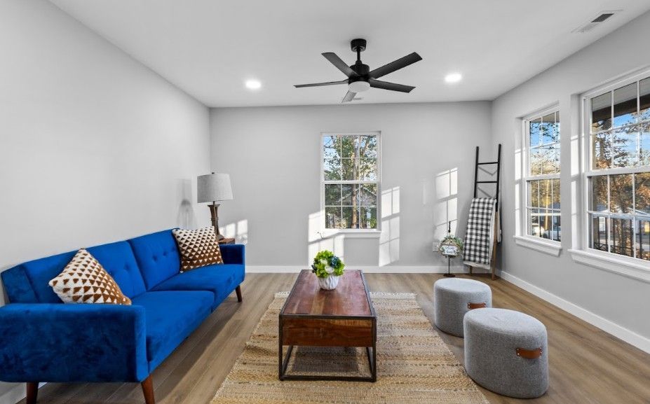 Bright modern living room with blue sofa, wood coffee table, gray ottomans, and large windows.