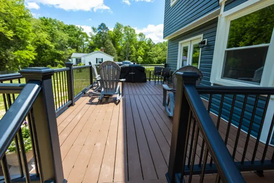 Wooden backyard deck with railing, chairs, grill, and a house side under a bright blue sky