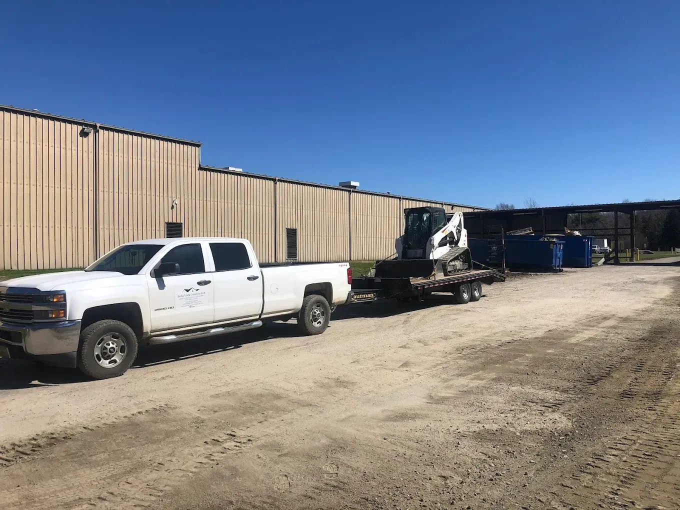 White pickup truck towing equipment in a dirt lot beside a tan industrial building
