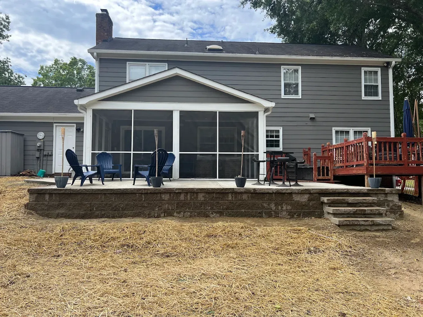 Backyard view of a gray house with screened porch, patio chairs, and a wooden deck over a stone retaining wall