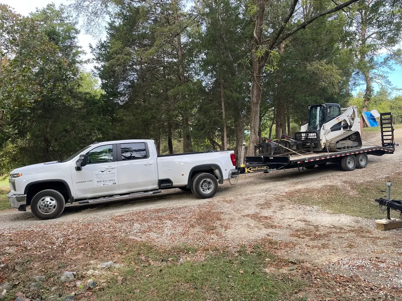 White pickup truck towing a trailer with a construction machine on a gravel driveway in a wooded area