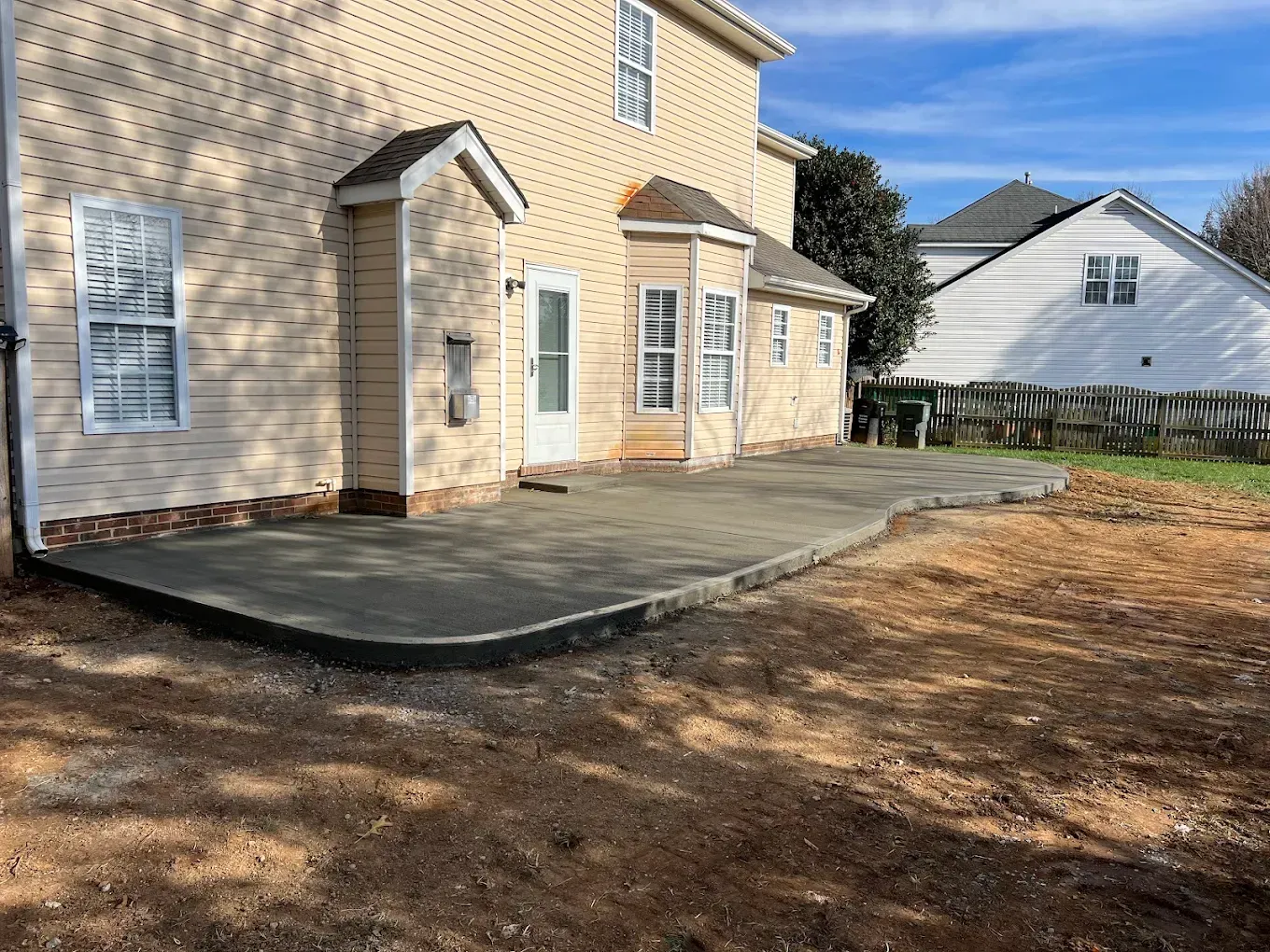 Backyard patio beside a beige house, with a concrete walkway and bare ground, next to a white neighboring home