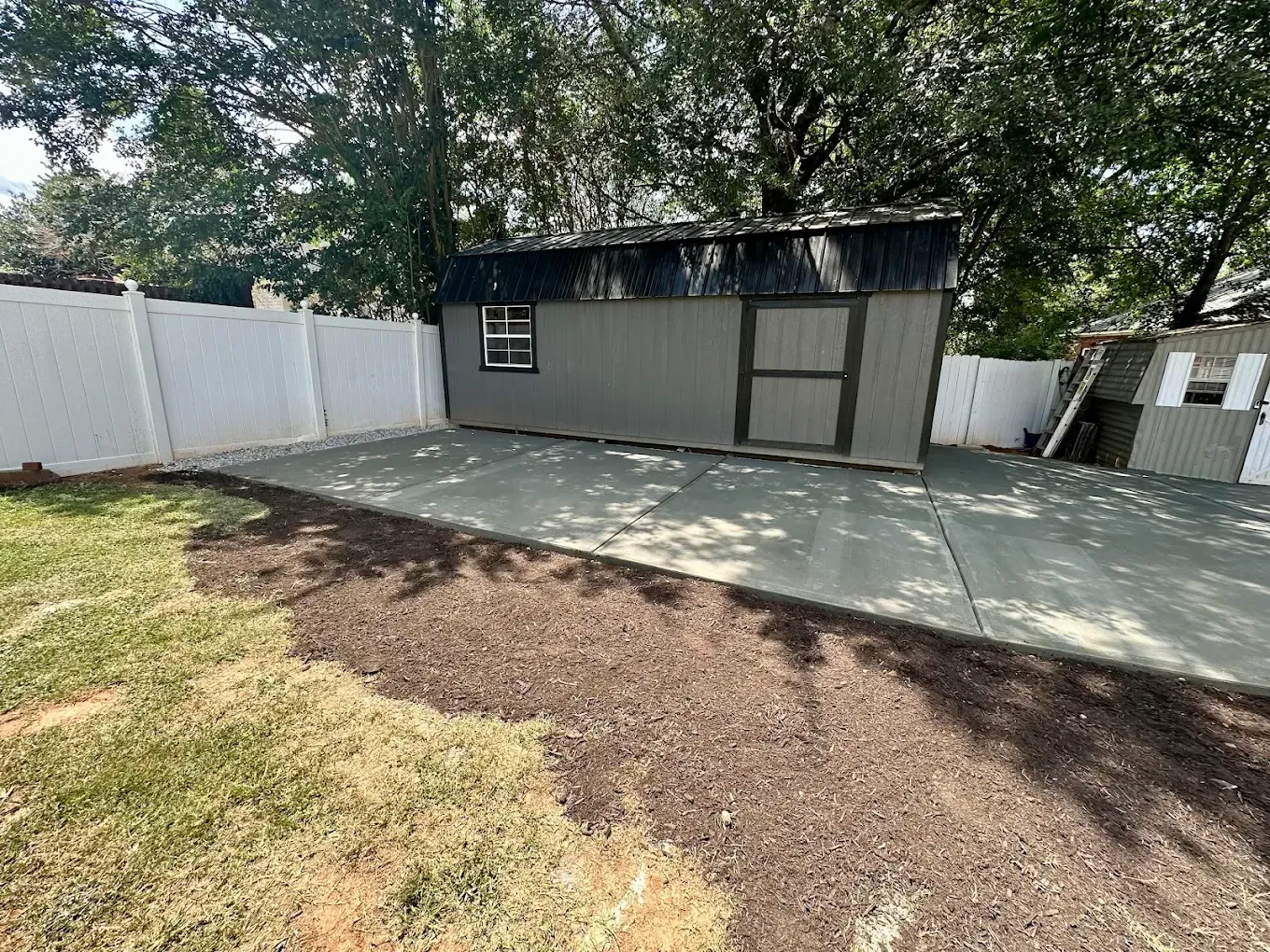 Backyard with gray shed, concrete patio, white fencing, and trees casting shade