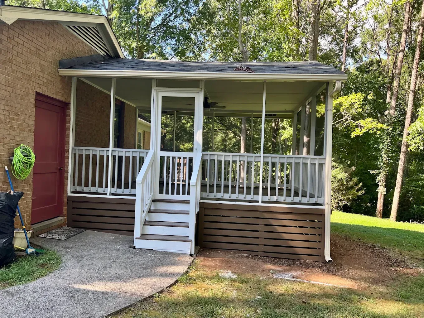 Small white porch with steps and railing attached to a brick house, surrounded by trees and grass.