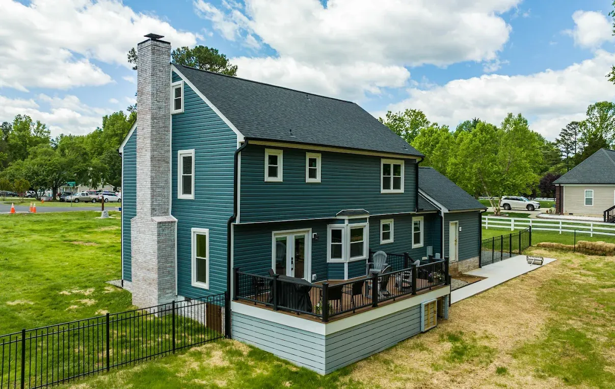 Blue two-story house with a porch, white trim, and stone chimney in a grassy yard under a cloudy sky