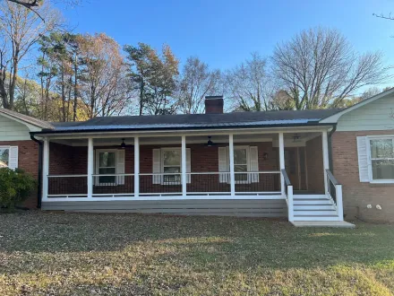 Front porch of a brick house with white railings, green shutters, and autumn trees in the background