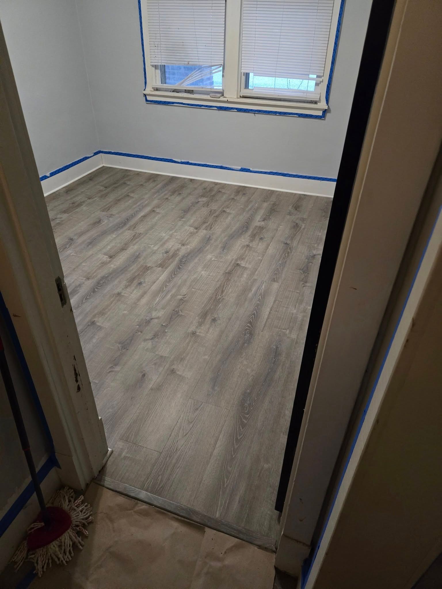 Room with grey flooring, window with blinds, and blue painter's tape along the walls.