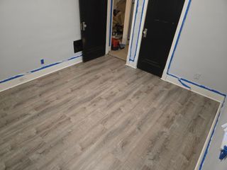 Room with gray-tone wood-look flooring, two black doors, and white walls with blue tape for painting.