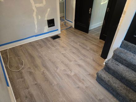 Newly installed wood-look flooring in a room with a black door, stairs, and blue painter's tape along the walls.