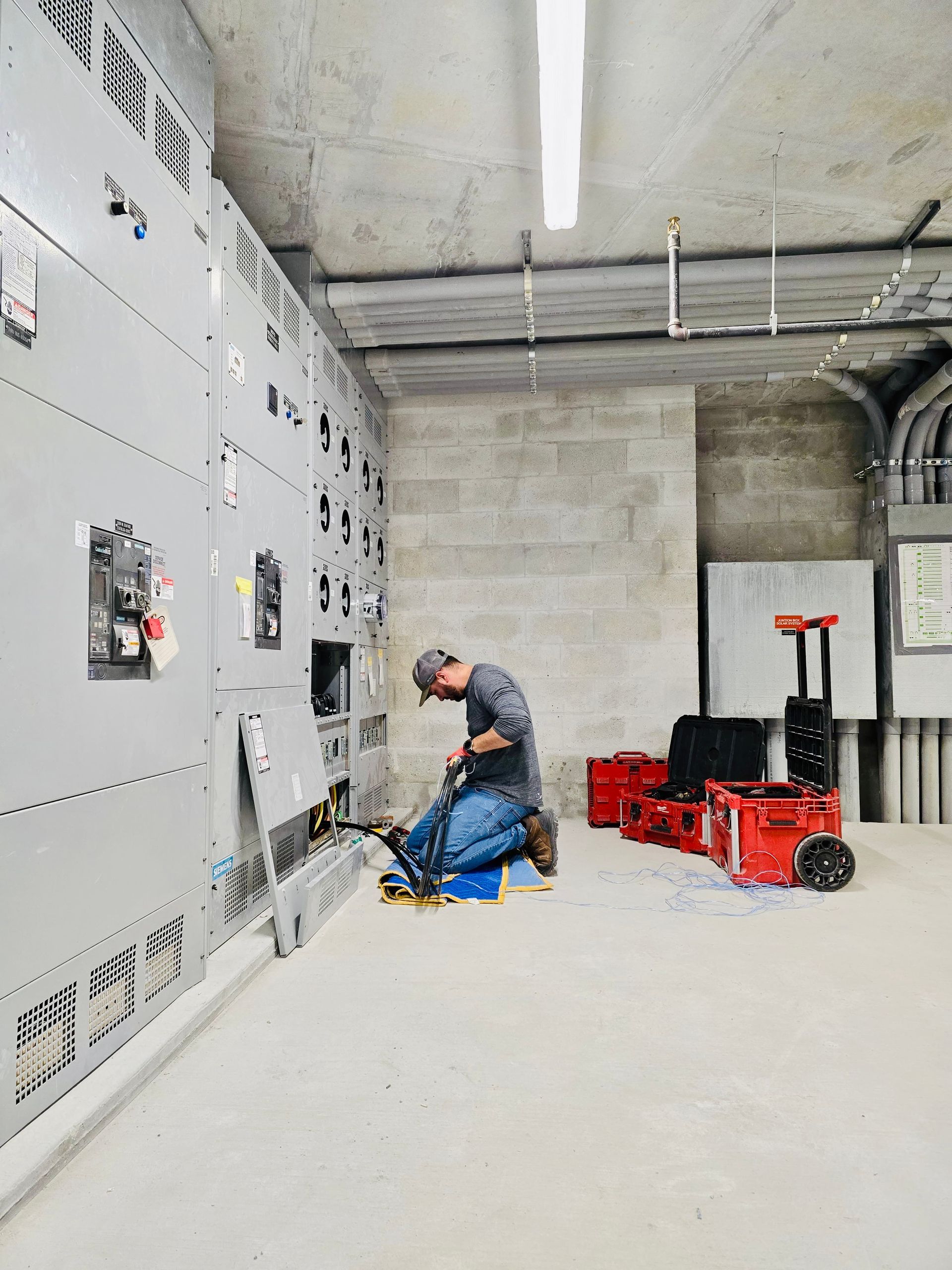 Technician kneels by electrical control panels in a concrete utility room with tools and equipment nearby.