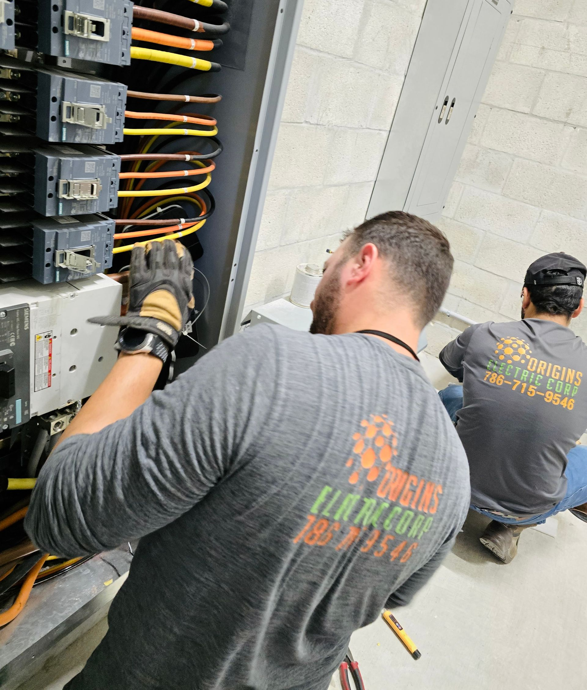 Technicians wiring electrical panels in a utility room, with exposed cables and equipment racks.