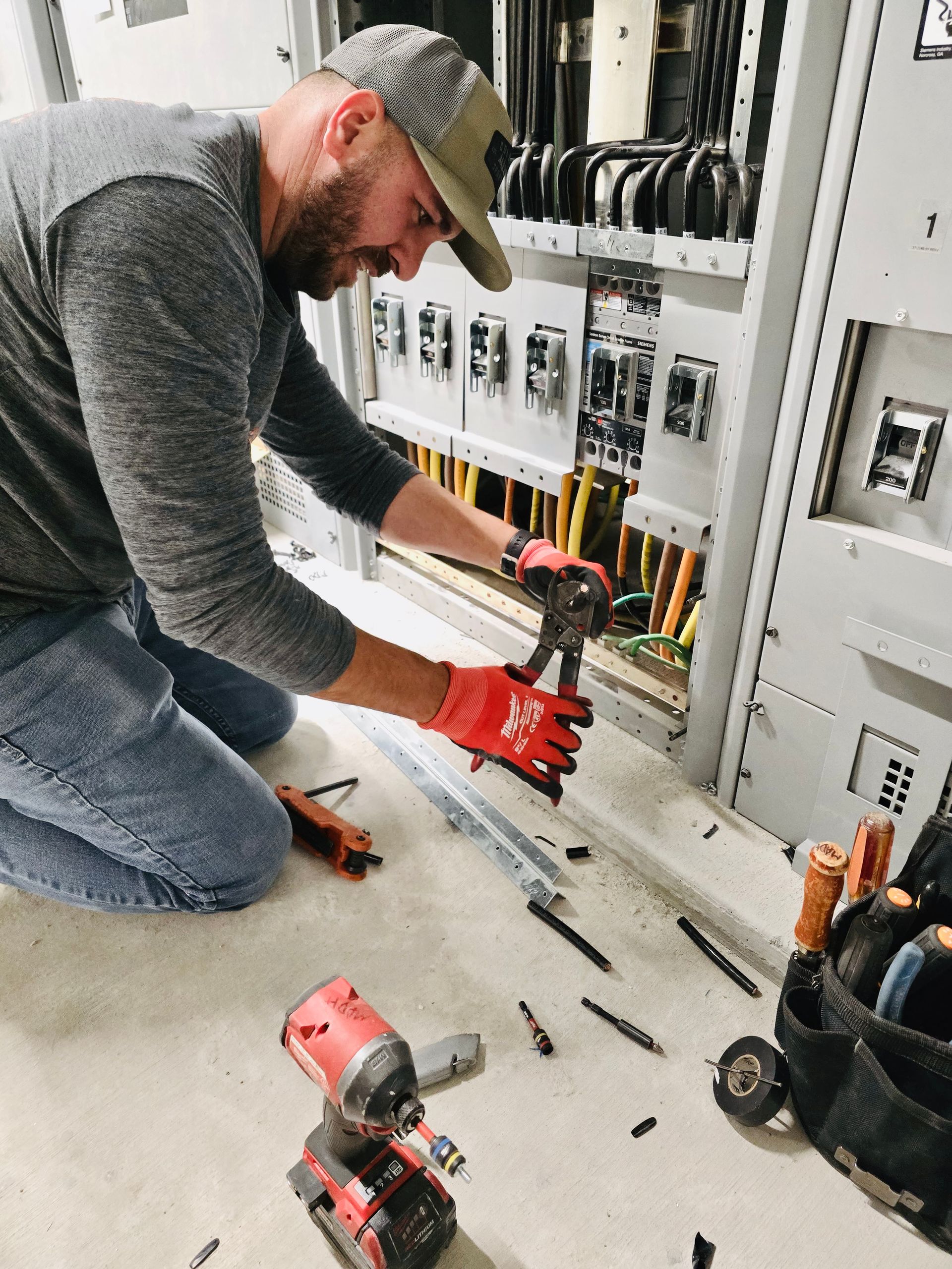 Technician kneeling at an open electrical panel, wiring cables with red gloves and tools nearby.