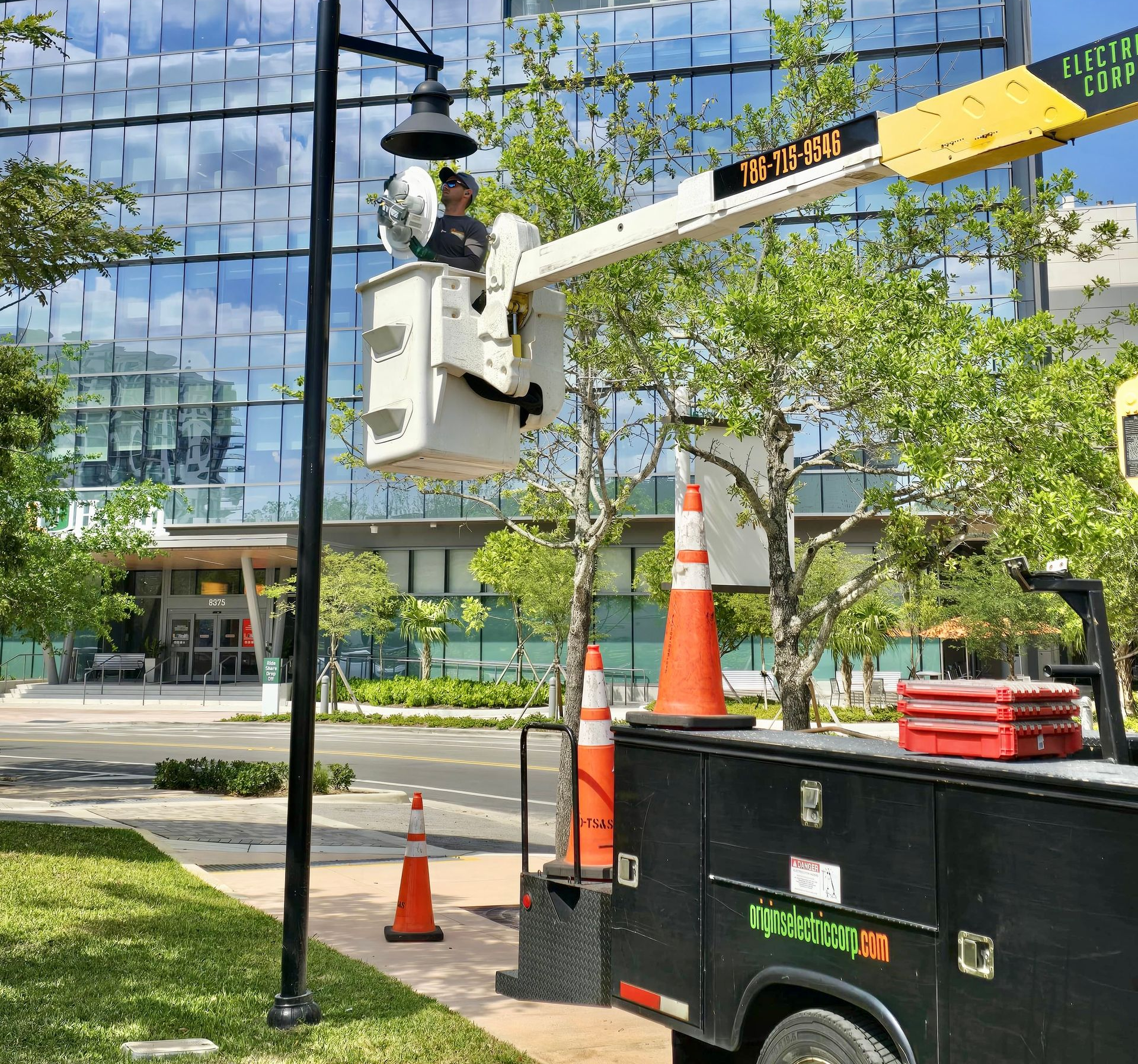 Utility worker in bucket truck trimming tree branches beside a glass office building