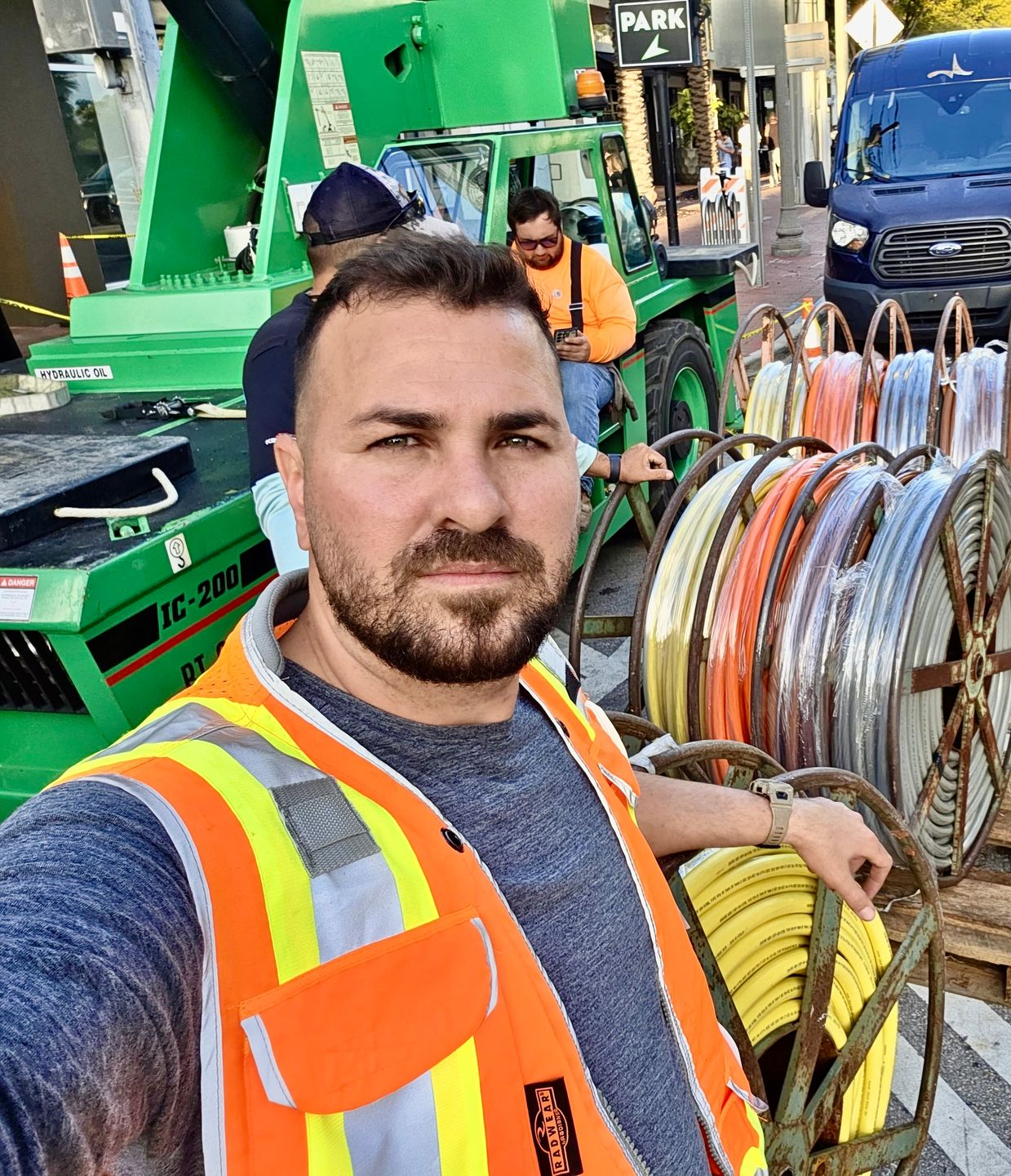 A person in a high-visibility vest stands in front of a green construction vehicle and several spools of colorful cable.