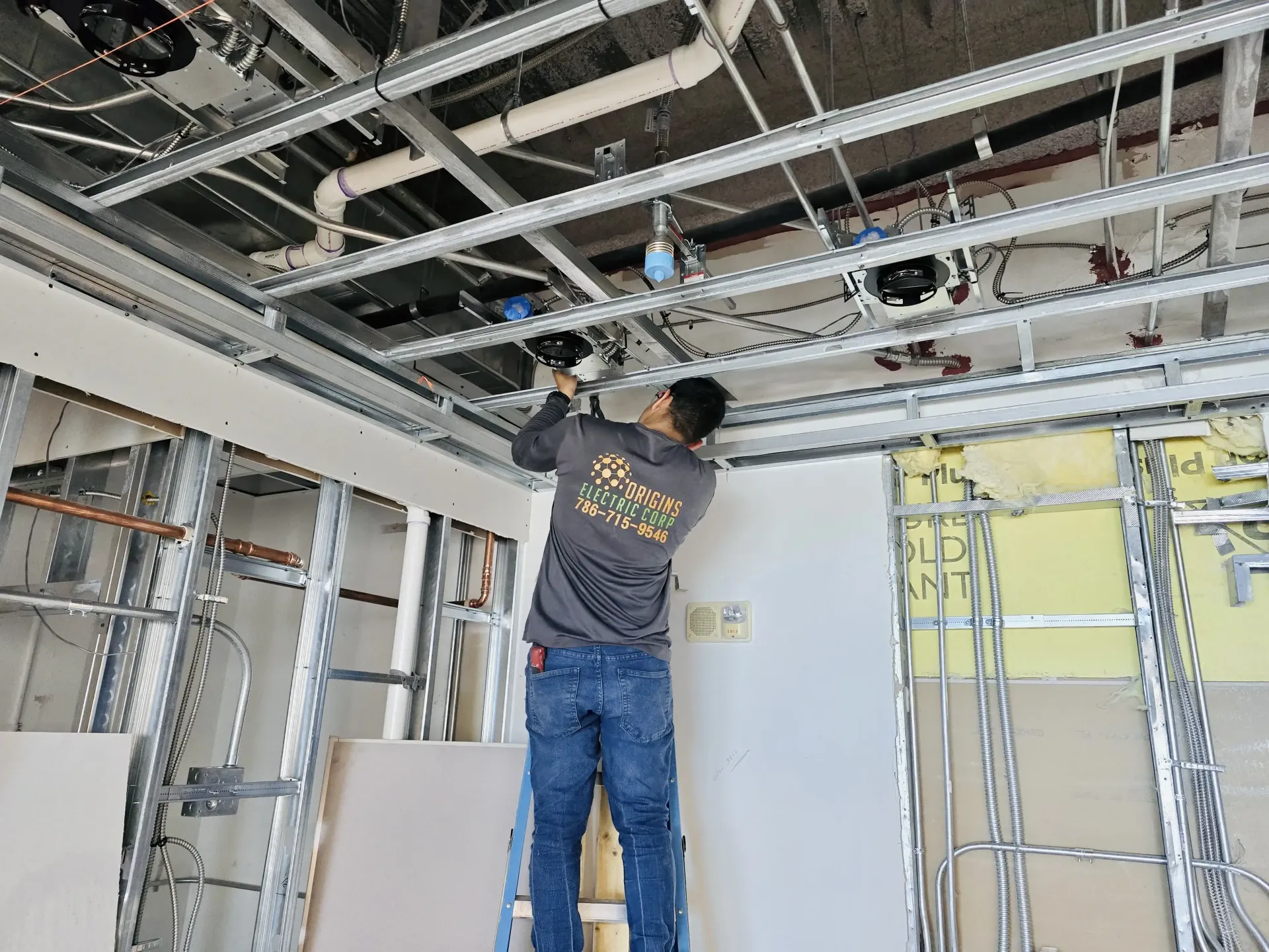 Worker installing overhead piping in an unfinished room with exposed ceiling beams and insulation.