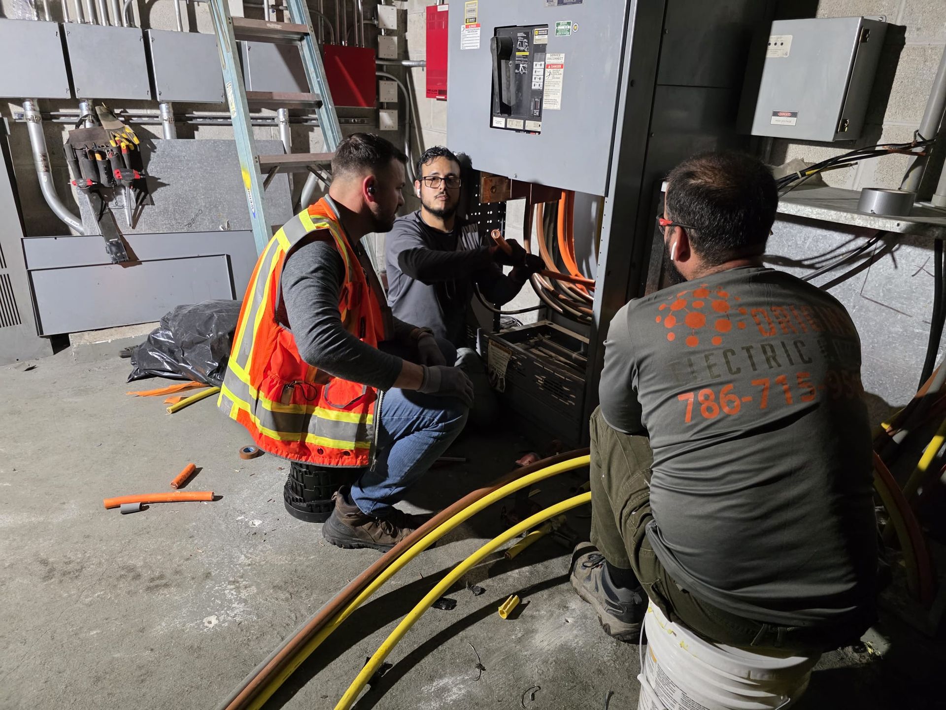 Workers in safety vests inspect electrical panels and cables inside an industrial room.