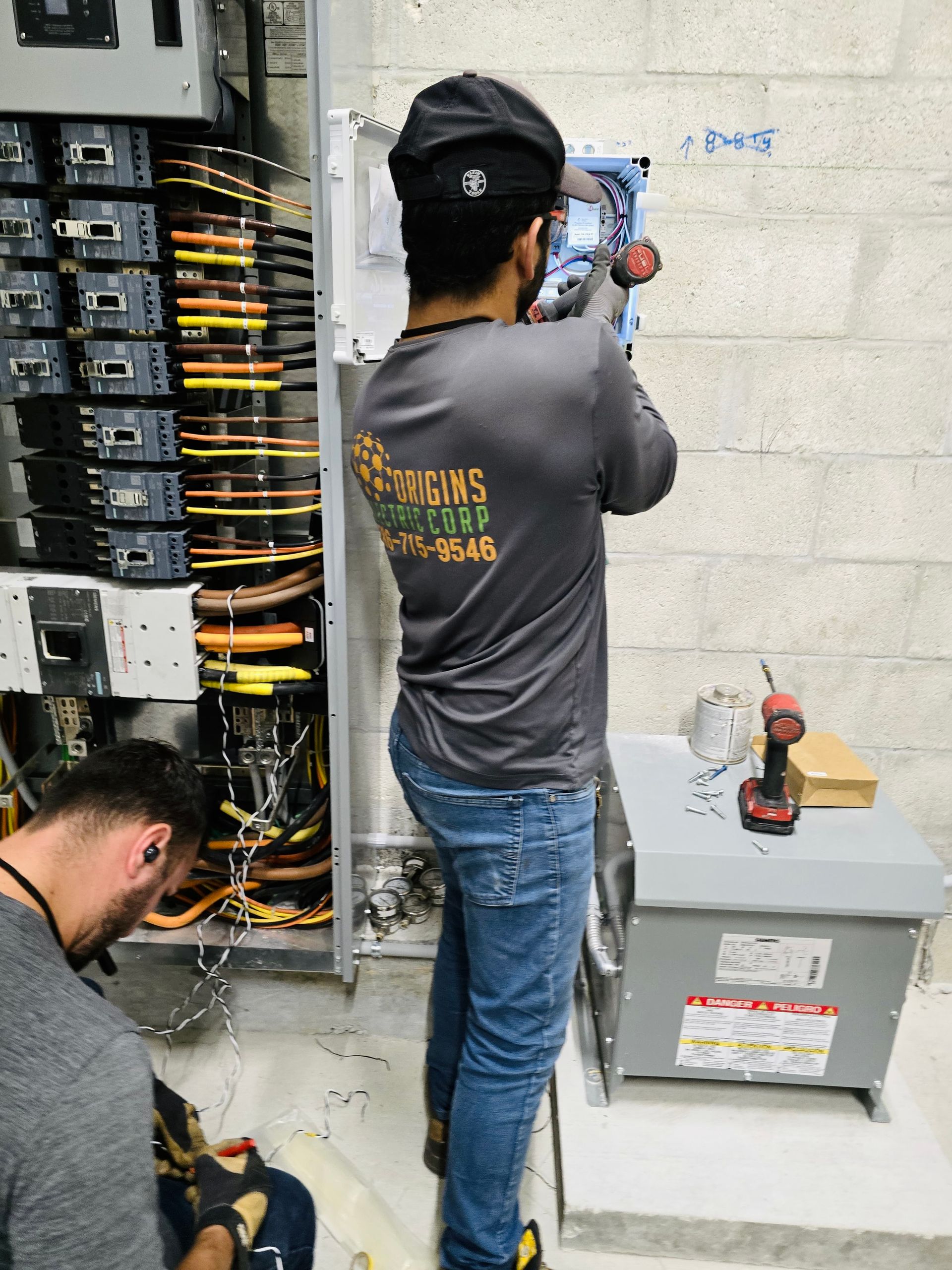 Technician inspecting electrical panel while another works on wiring in a utility room