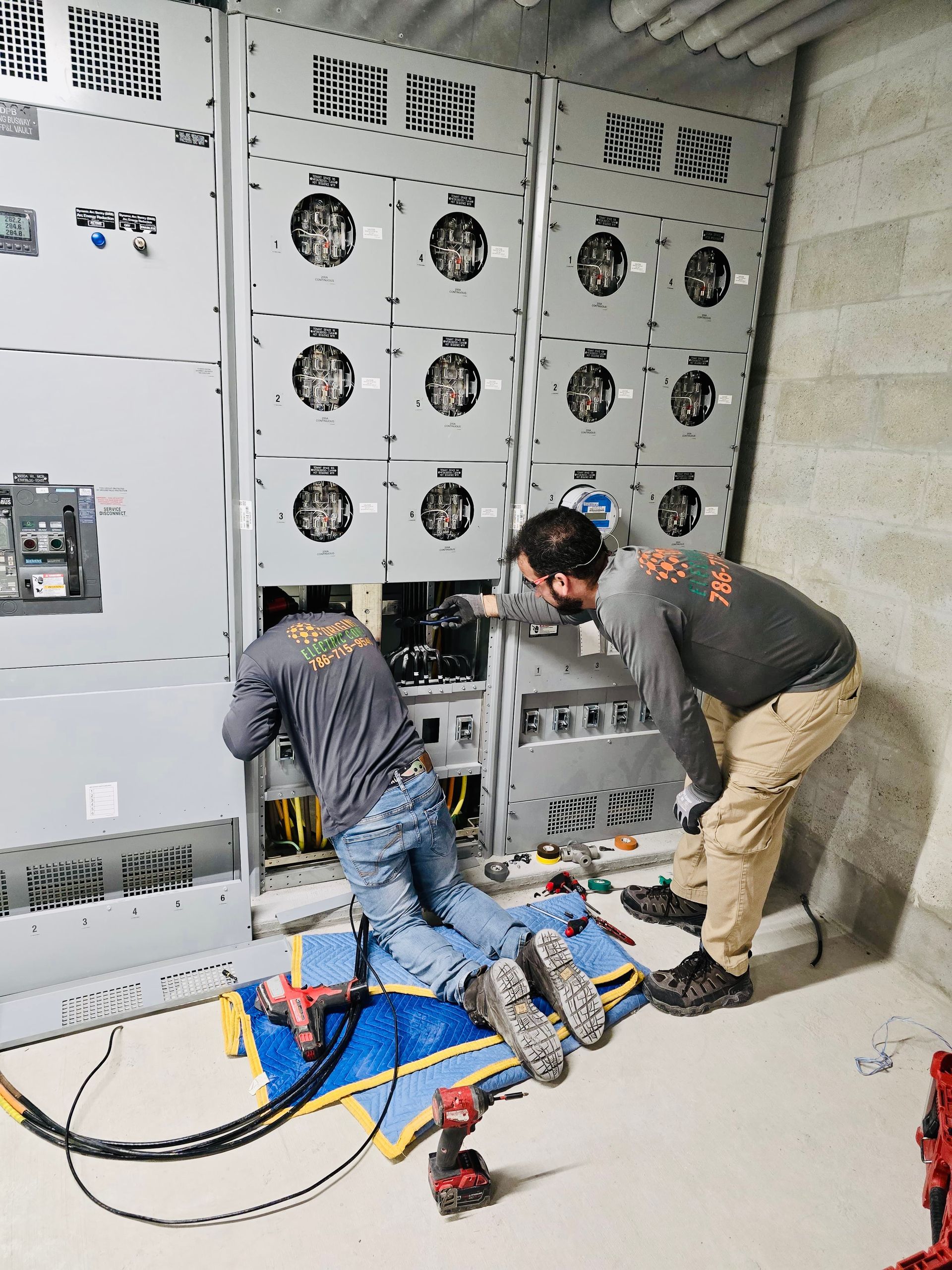 Two workers servicing electrical panels in a utility room, with cables and tools on the floor.