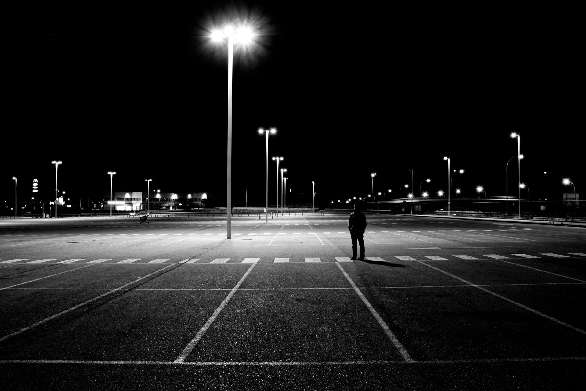 A lone figure stands in the center of a vast, dimly lit parking lot at night under bright overhead pole lights.