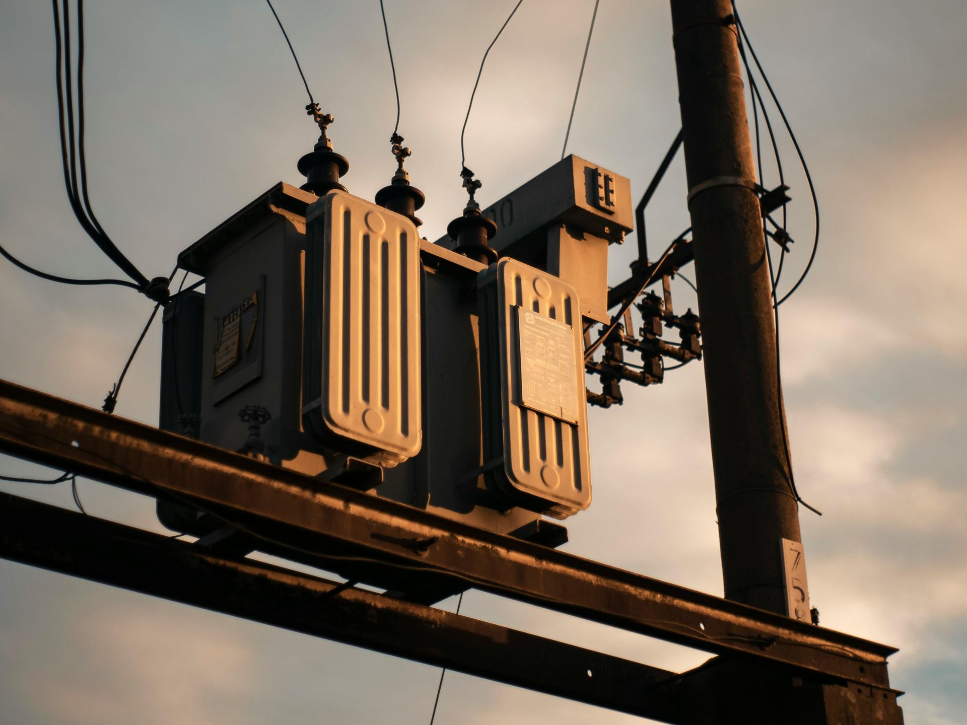 A gray electrical transformer mounted on a utility pole against a cloudy sky at sunset.