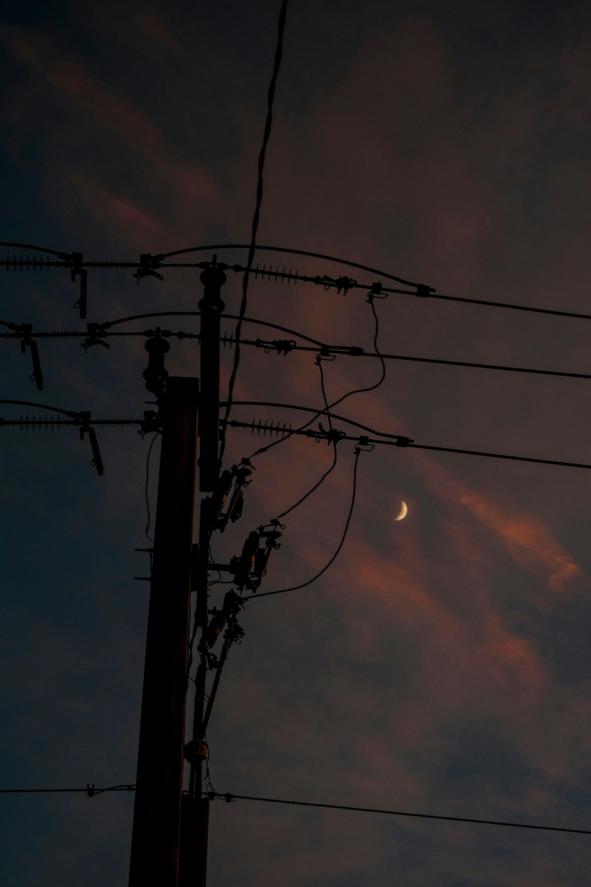 A silhouette of a utility pole and power lines against a twilight sky with a visible crescent moon.