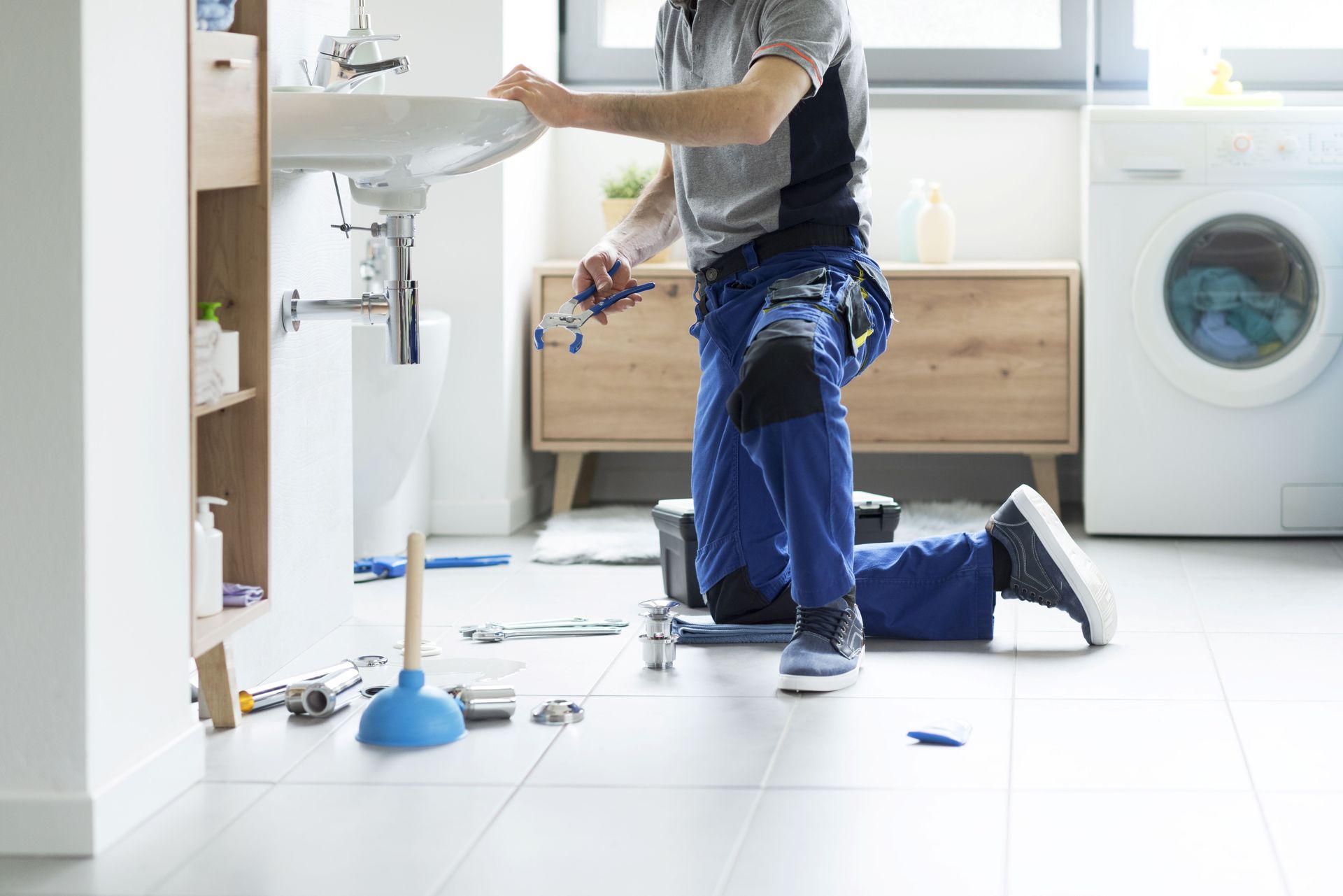 A plumber kneels fixing a sink, tools scattered, the bathroom lit by window light. A plumber kneels fixing a sink, tools scattered, the bathroom lit by window light.