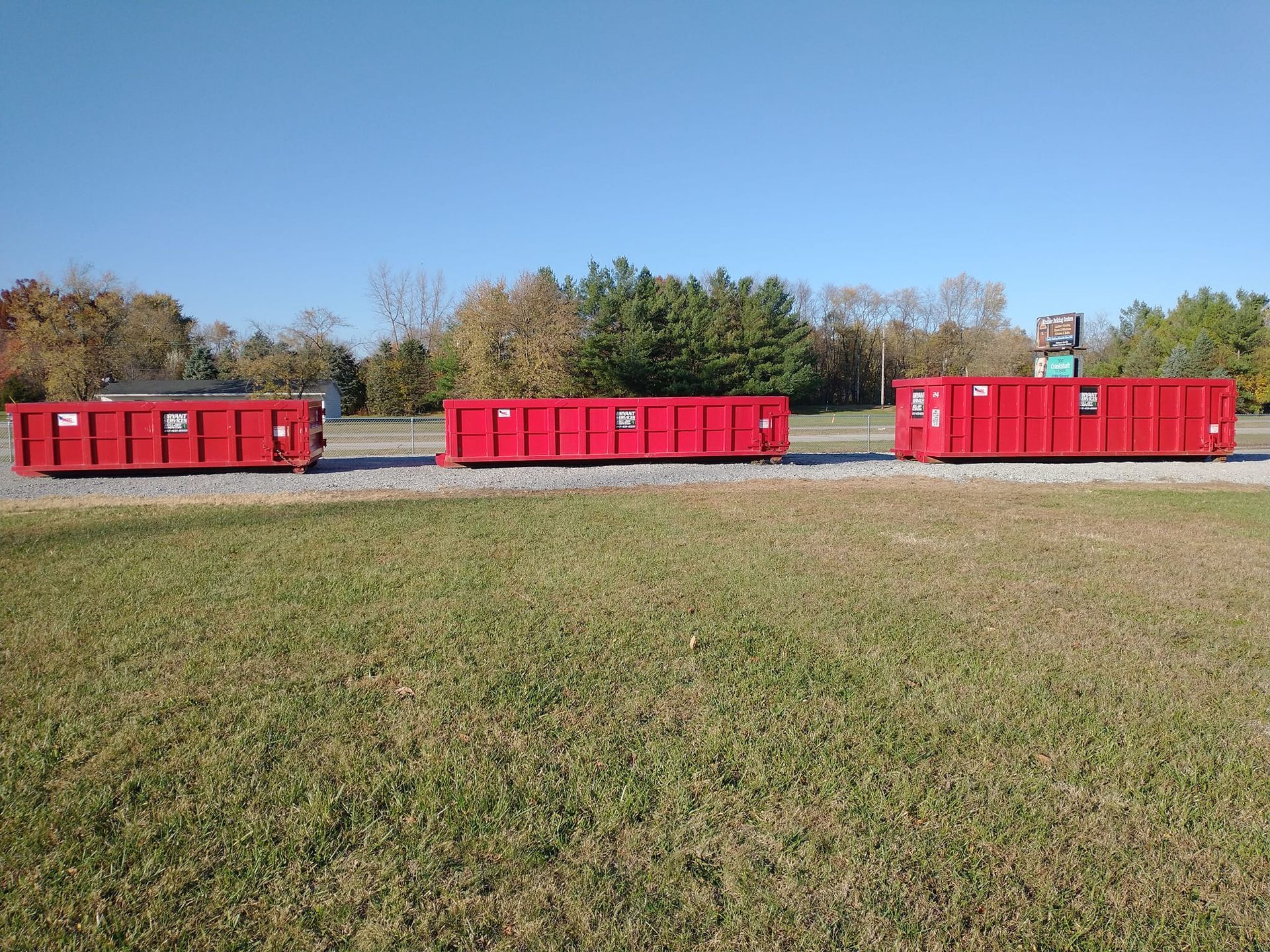 Three bright red roll-off dumpsters sit in a row on a gravel lot under a clear blue sky.