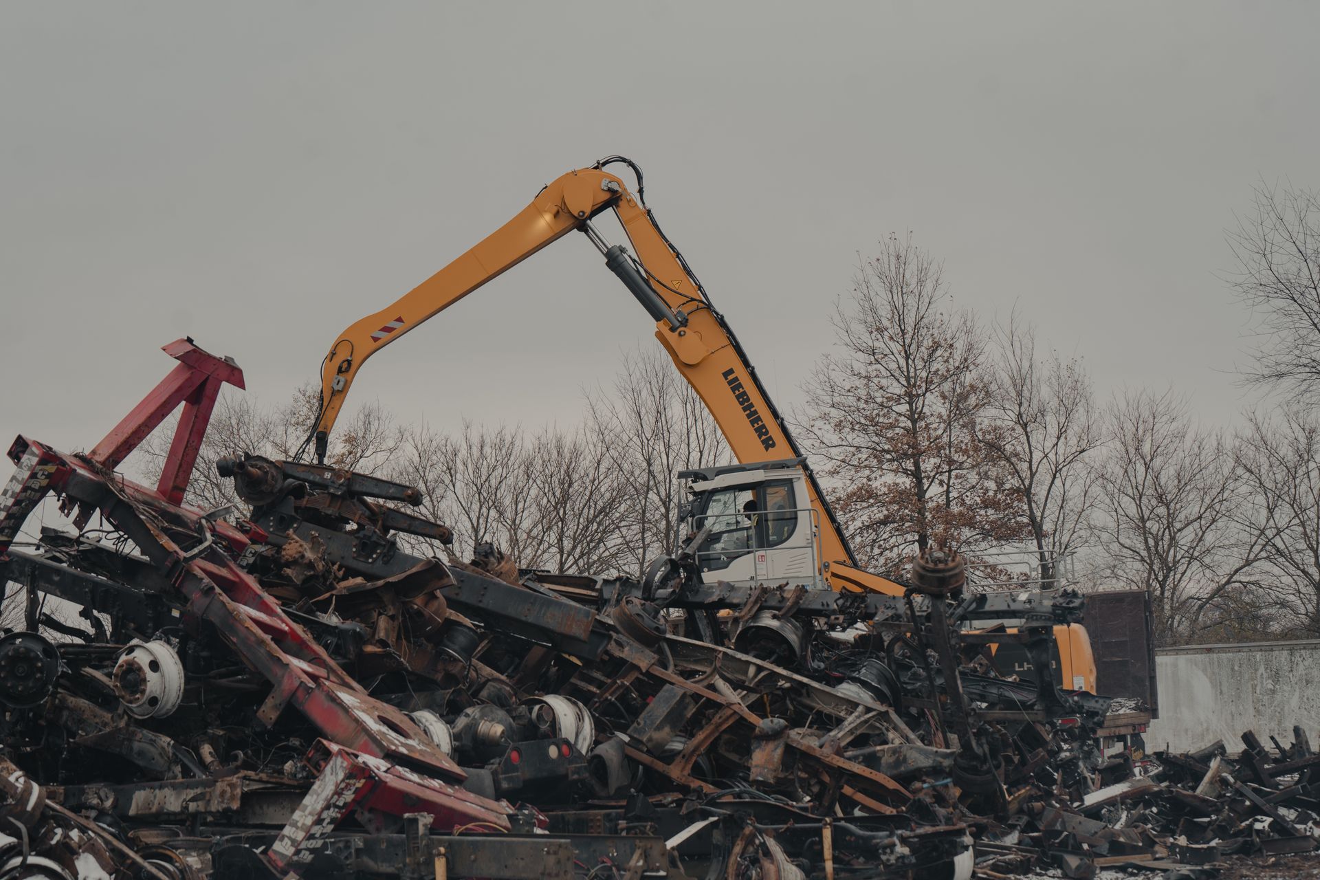 A yellow excavator operates on a large, messy pile of dark metal scrap and debris outdoors under a grey sky.