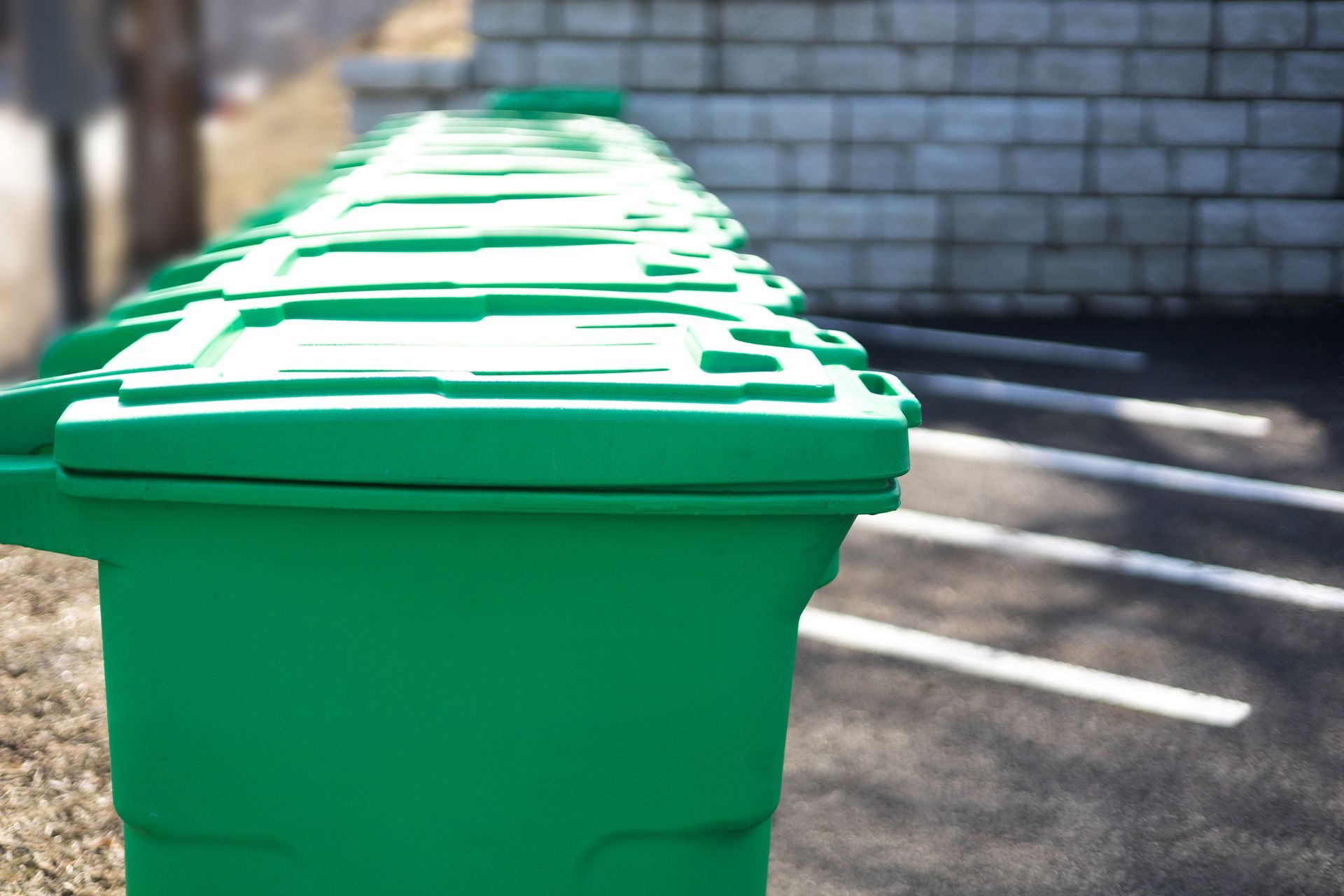 A row of identical green plastic trash bins lined up next to a parking lot and a brick wall.