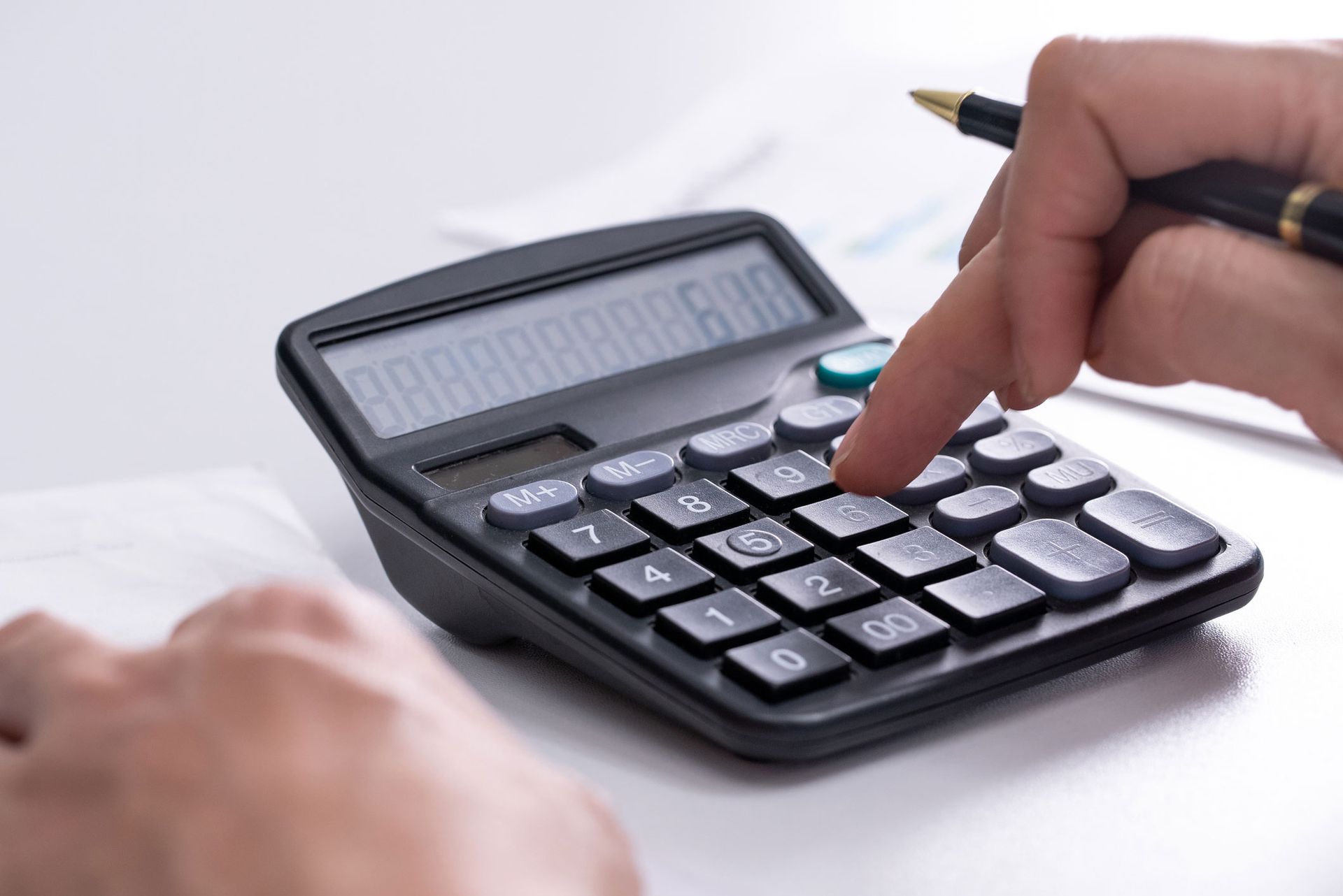 A person uses a black calculator on a white surface, with a hand pressing a key while holding a pen.