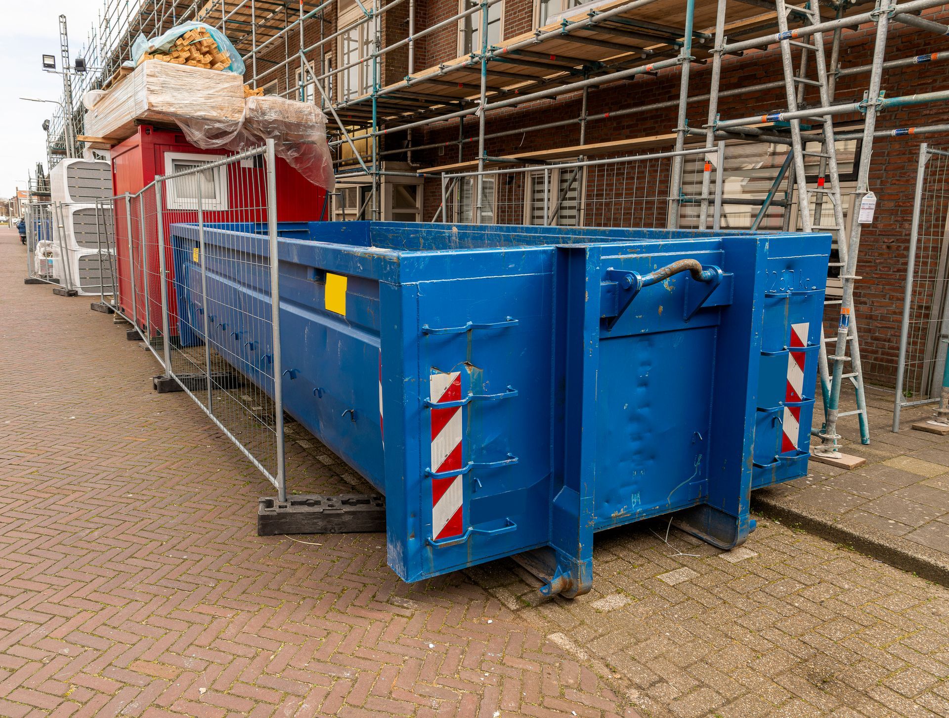 A large blue dumpster sits on a brick-paved street next to scaffolding and a small red construction site office.