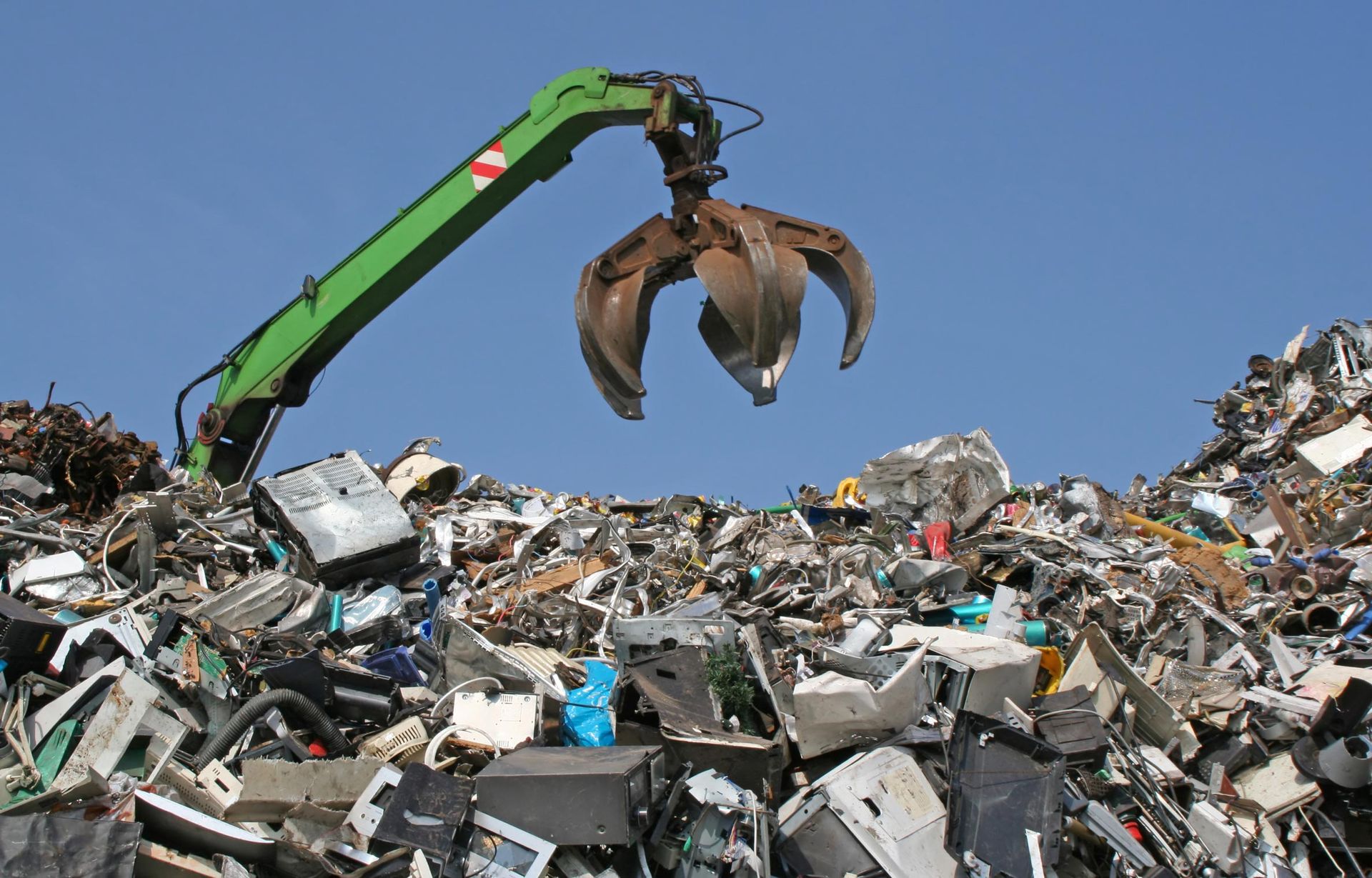 A green mechanical grabber crane positioned over a large pile of electronic waste and scrap metal against a blue sky.