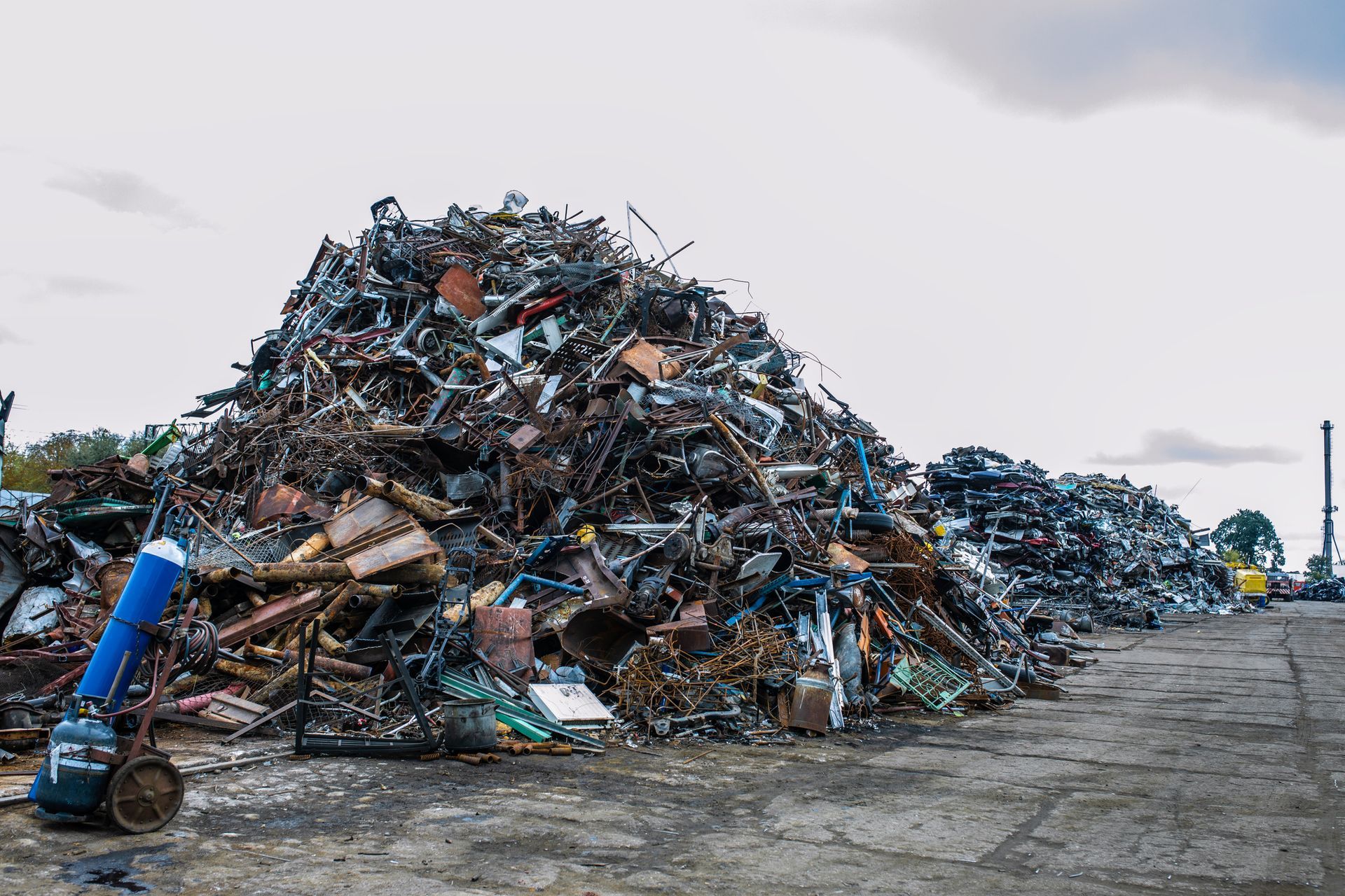 Piles of metal scrap tower under open sky, showcasing quick and reliable metal recycling. A large, sprawling pile of scrap metal and industrial waste sits on an outdoor concrete lot under a cloudy sky.