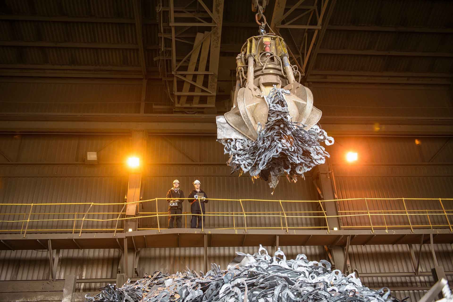 Two people in hard hats stand on a factory platform watching a large crane claw lift a pile of metal scrap.