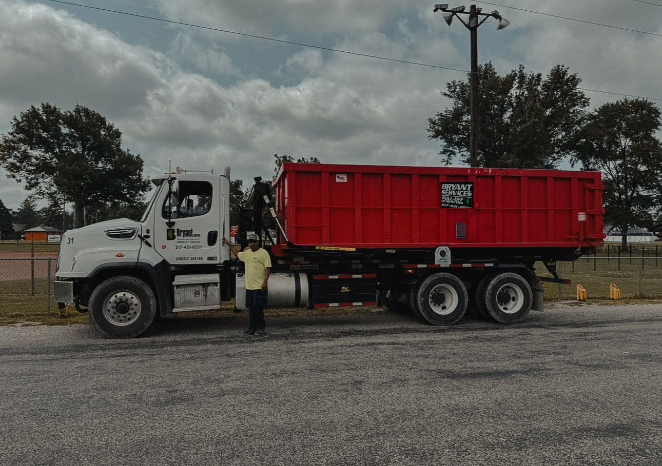 A person stands next to a white roll-off truck with a large red dumpster, parked on a gravel surface under a cloudy sky.