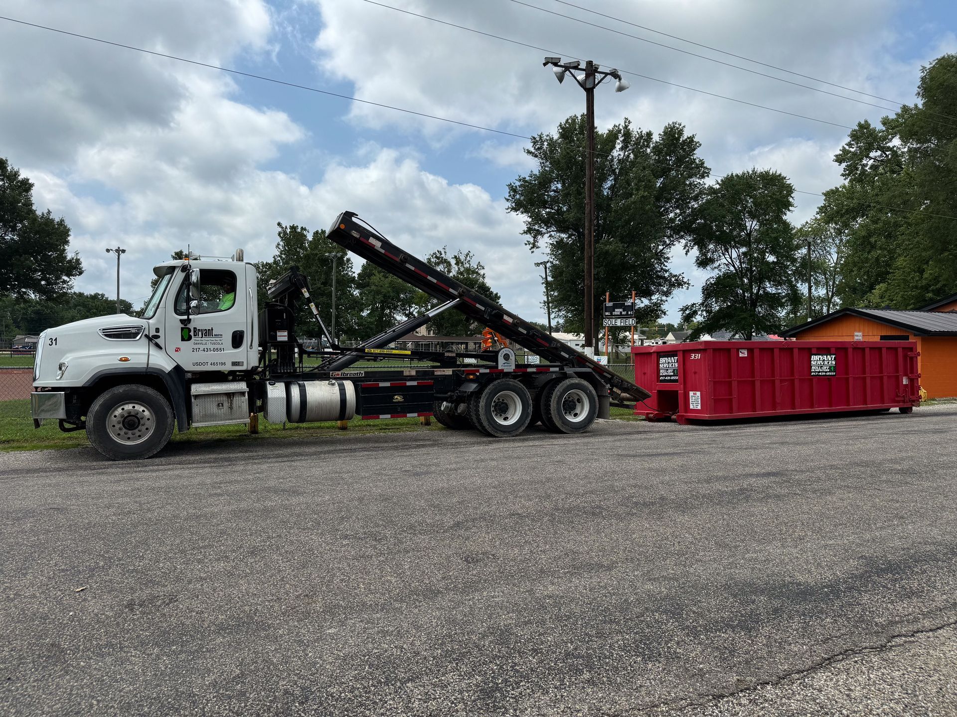 A white roll-off truck positioning a large red dumpster on a gravel lot under a cloudy sky.