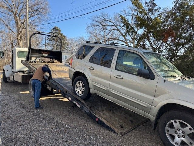 A person uses a tow truck to load a light-colored SUV onto the tilted flatbed trailer outdoors.