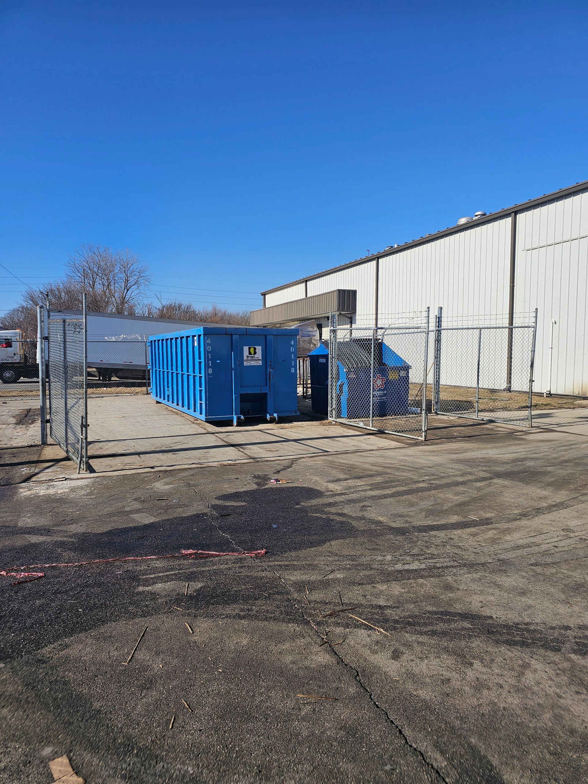Two blue industrial dumpsters sit on an asphalt lot beside a large warehouse and chain-link fence under a clear blue sky.