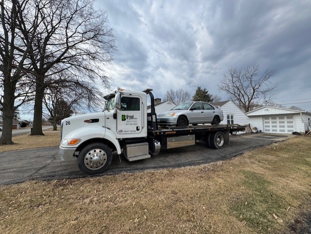 A white tow truck carrying a silver car parked on a paved driveway in front of a house on a cloudy day.