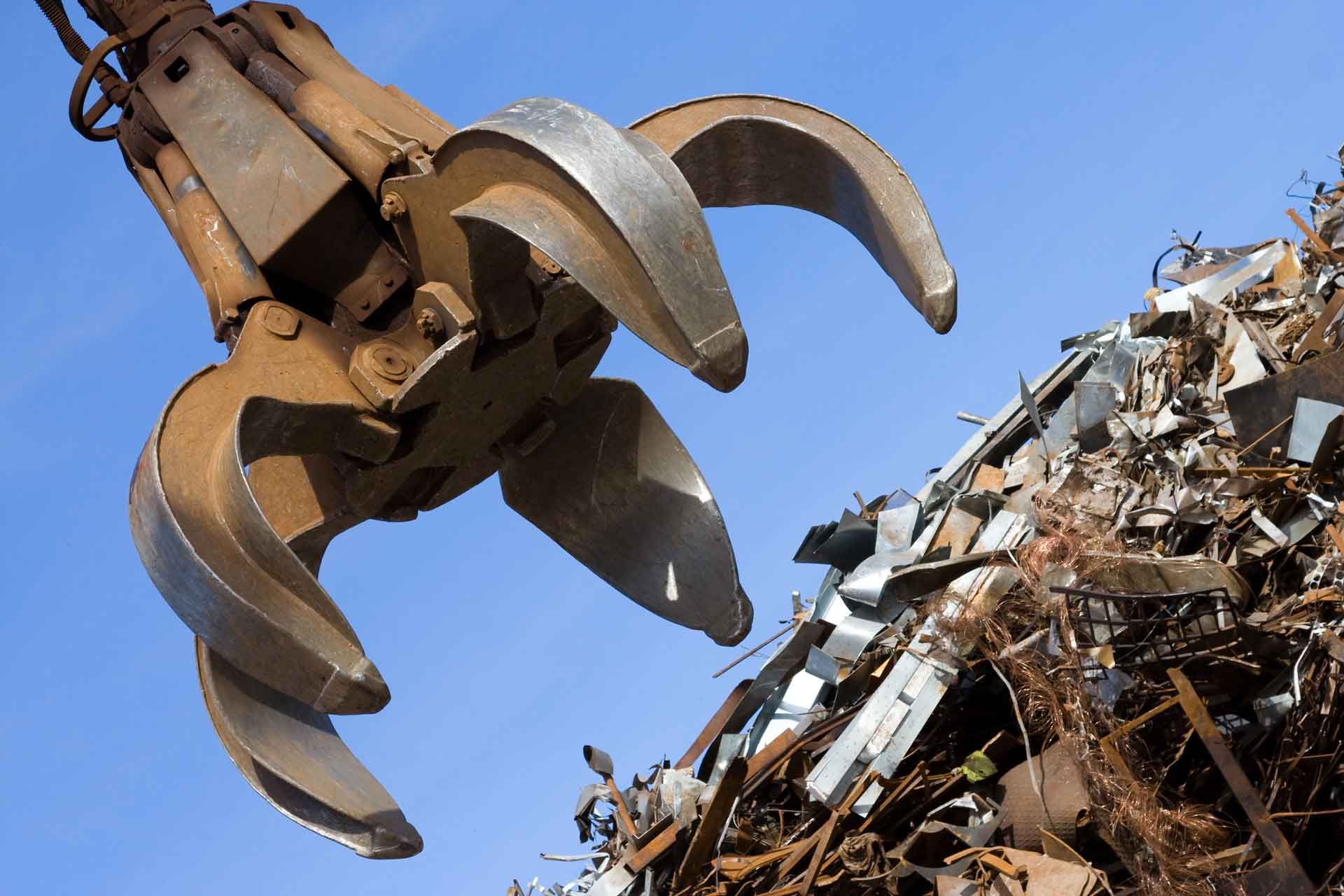 A metal scrap grapple hanging against a blue sky, positioned above a large pile of industrial metal debris.