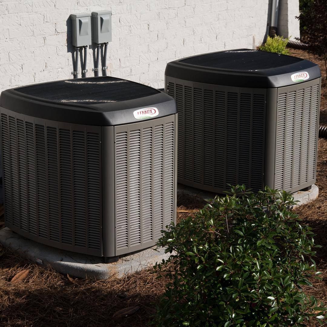 Two outdoor air conditioning units beside a house wall and shrubs