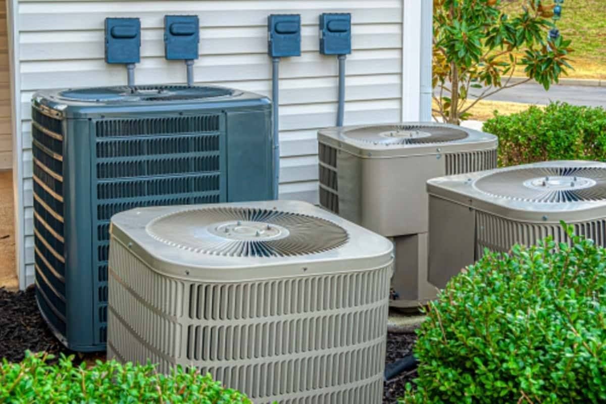 Outdoor HVAC units beside a house, with blue and beige air conditioners near green shrubs