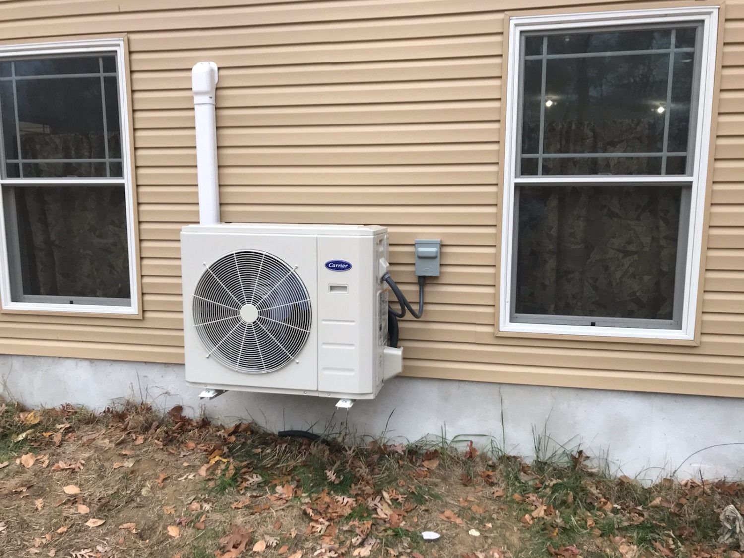 Outdoor HVAC unit mounted on tan house siding beneath two windows
