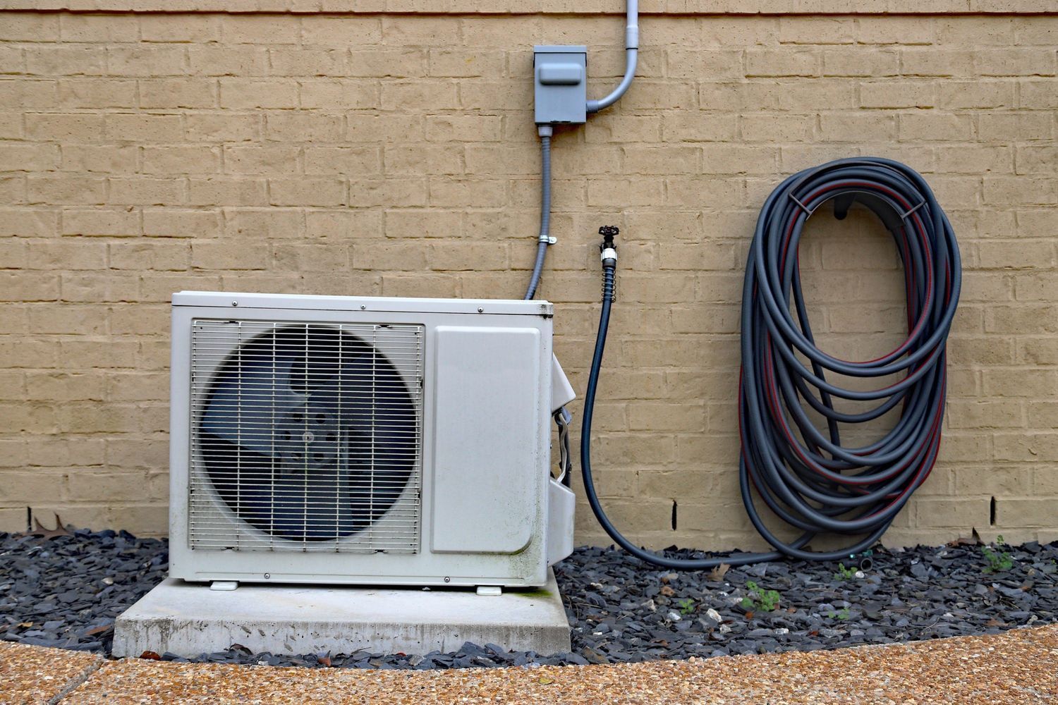 Outdoor air conditioner unit beside a beige brick wall with coiled hoses and an electrical box