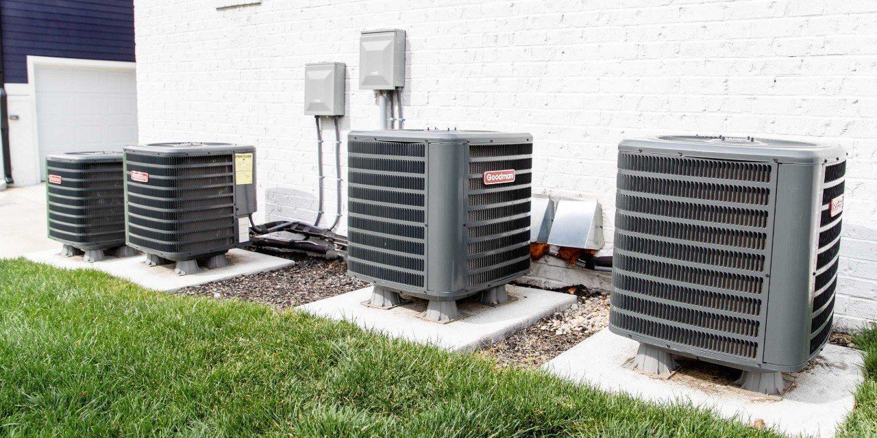 Row of outdoor air conditioning units beside a building, with grass in front and gravel around them.