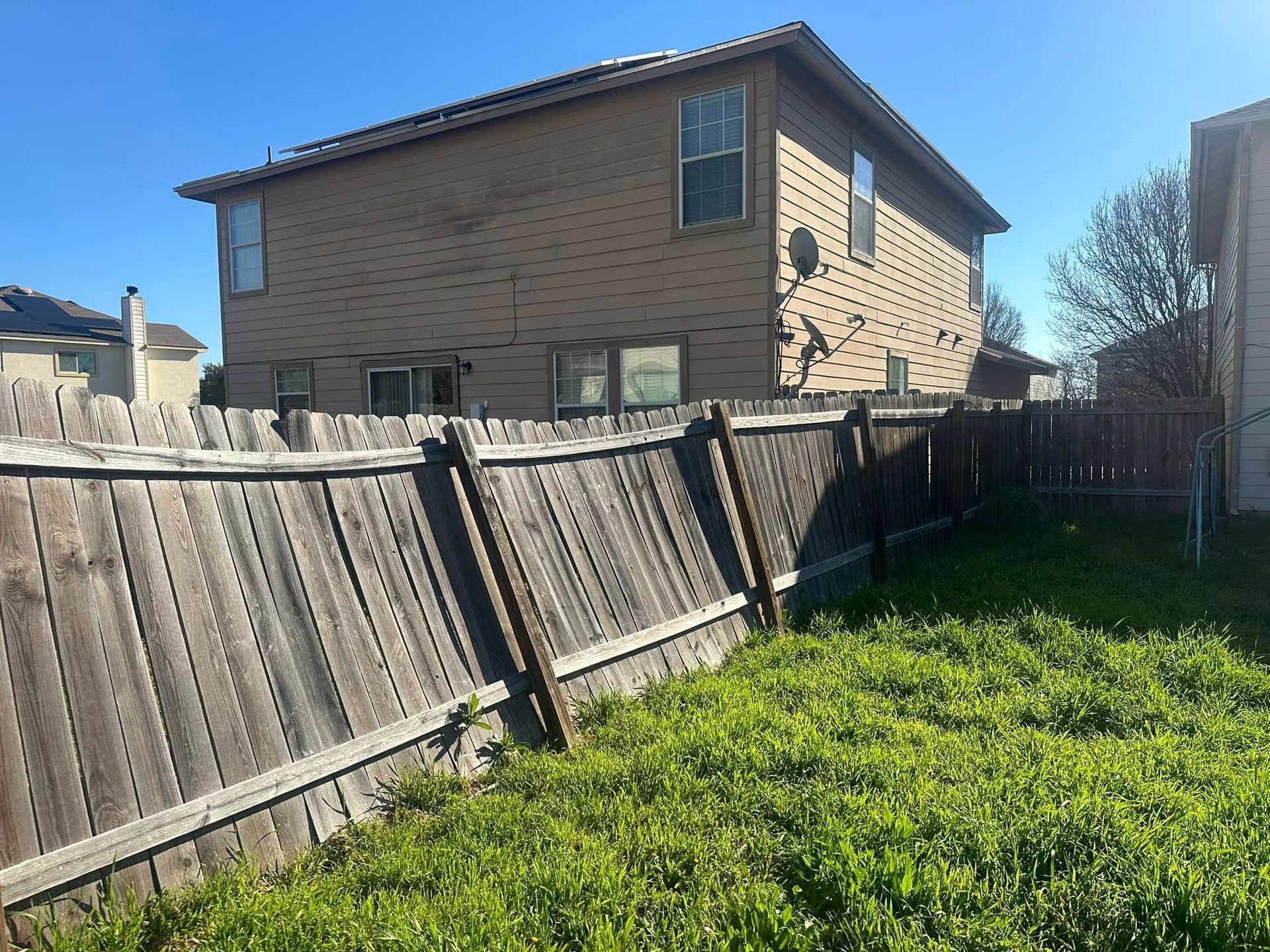 Backyard with leaning wooden fence, green grass, and a two-story brick house under a blue sky