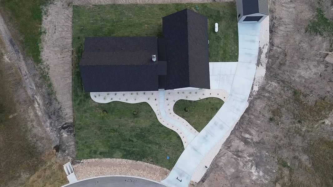 Aerial view of a house with a dark roof, light walkway, and surrounding grass and dirt.